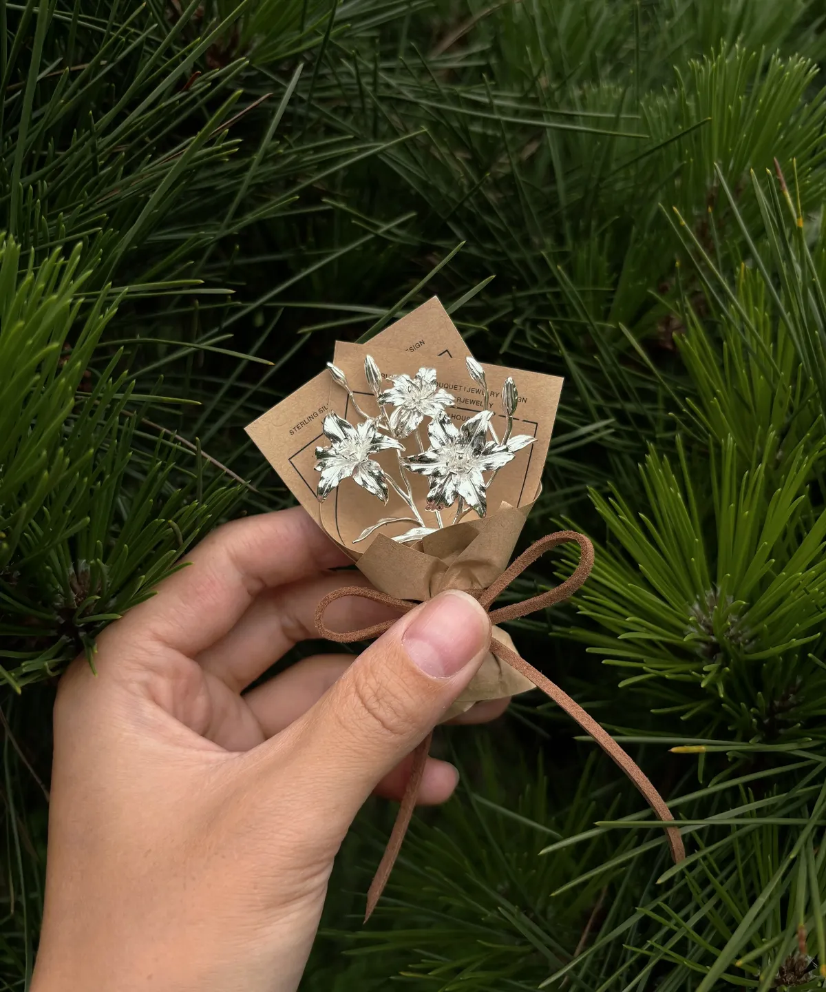 Hand holding a delicate mini lily bouquet wrapped in brown paper with two small cards, set against lush green pine needles. Perfect for Lazesoul handmade art gifts, 925 sterling silver Lily Bouquet jewelry and keepsakes.