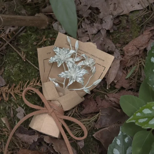A 925 sterling silver mini lily bouquet wrapped in rustic brown paper, tied with twine, resting on a forest floor. Handmade by Lazesoul, this elegant Lily Bouquet is a timeless art gift blending nature-inspired beauty with artisanal craftsmanship.