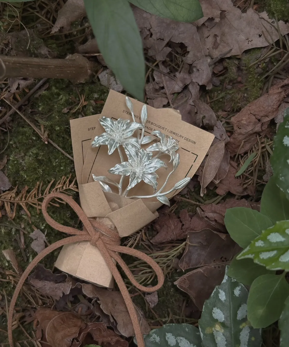 A 925 sterling silver mini lily bouquet wrapped in rustic brown paper, tied with twine, resting on a forest floor. Handmade by Lazesoul, this elegant Lily Bouquet is a timeless art gift blending nature-inspired beauty with artisanal craftsmanship.