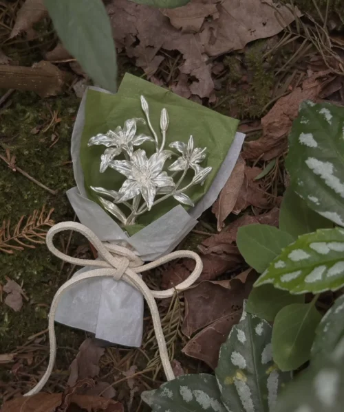 Handmade 925 sterling silver mini lily bouquet wrapped in green and white paper, tied with a white bow, resting on a forest floor. Elegant art gift blending metallic floral beauty with natural woodland charm. Perfect for Lily Bouquet lovers.