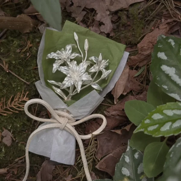 Handmade 925 sterling silver mini lily bouquet wrapped in green and white paper, tied with a white bow, resting on a forest floor. Elegant art gift blending metallic floral beauty with natural woodland charm. Perfect for Lily Bouquet lovers.