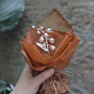 Hand holding a rustic paper-wrapped 925 sterling silver Lily of the Valley Bouquet, handmade by Lazesoul. Elegant bell-shaped flowers and leaves create a timeless art gift, blending nature-inspired beauty with artisanal craftsmanship.