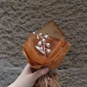 Hand holding a 925 sterling silver mini Lily Of The Valley Bouquet wrapped in rustic brown paper with twine, Lazesoul Handmade Art Gifts. Elegant metallic flowers against a stone wall, perfect for unique art gifts and nature-inspired jewelry lovers.