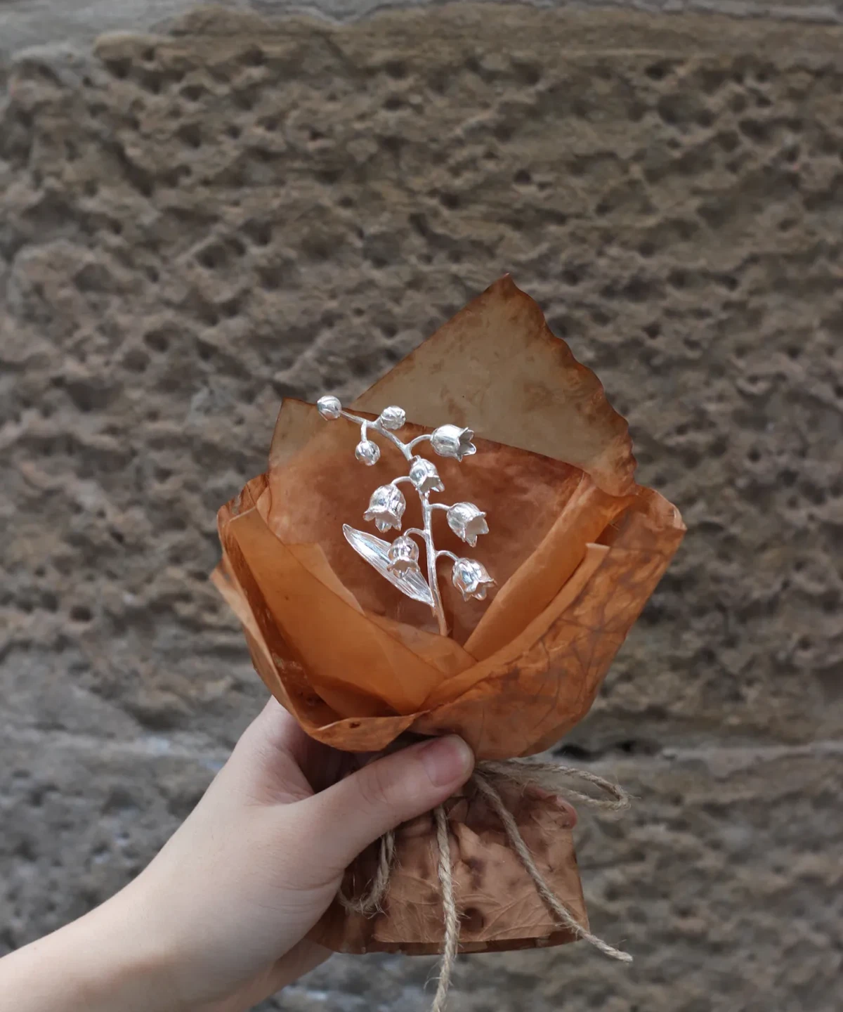 Hand holding a 925 sterling silver mini Lily Of The Valley Bouquet wrapped in rustic brown paper with twine, Lazesoul Handmade Art Gifts. Elegant metallic flowers against a stone wall, perfect for unique art gifts and nature-inspired jewelry lovers.