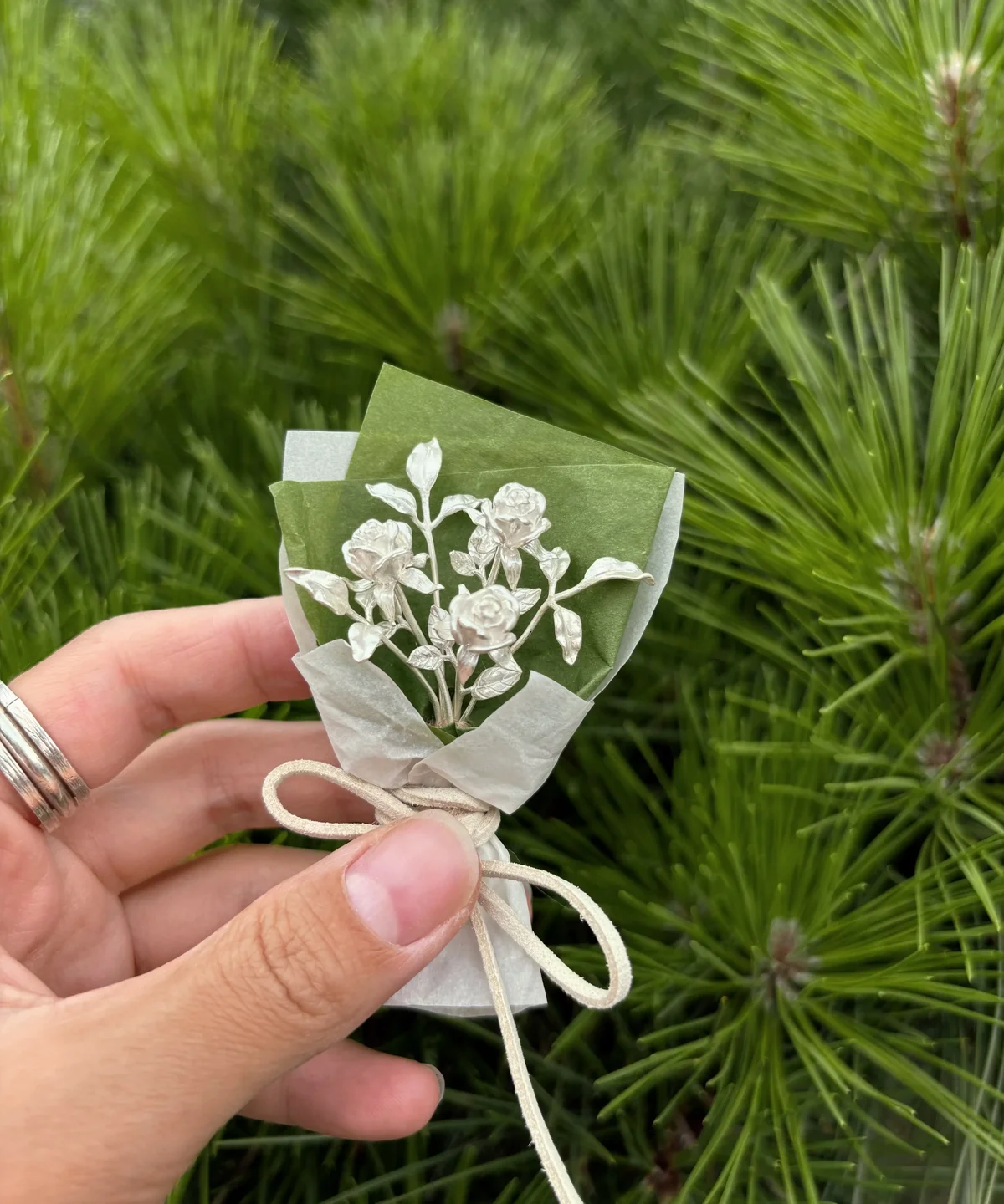 Hand holding a 925 sterling silver mini rose bouquet wrapped in green and white paper with a beige bow, Lazesoul handmade art gifts, nature-inspired jewelry and decorative keepsake, perfect rose bouquet for unique art gifts lovers.