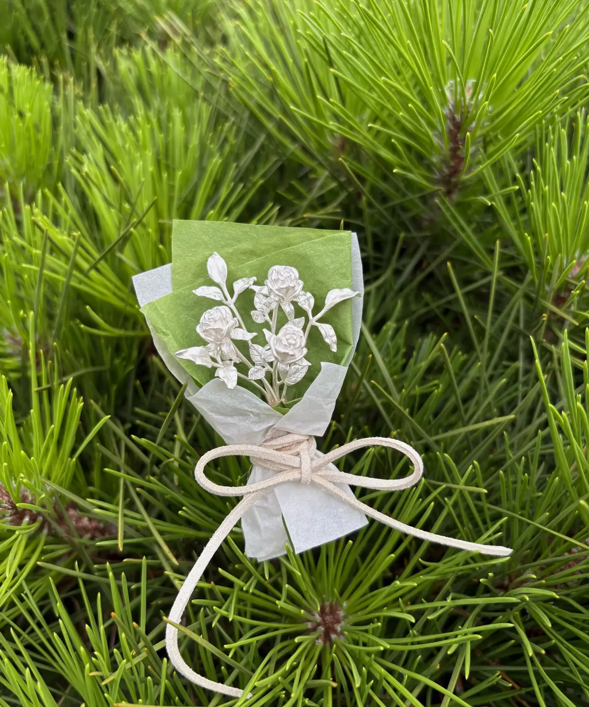 A delicate 925 sterling silver mini rose bouquet, handcrafted with intricate detail, wrapped in green and white paper, resting on pine needles. Perfect handmade art gifts for nature lovers and collectors of unique rose bouquet designs.