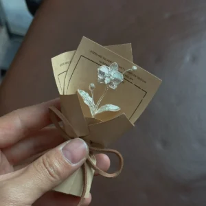 Hand holding a 925 sterling silver Phalaenopsis mini bouquet art gift, wrapped in brown paper with suede tie, featuring two delicate handmade silver flower pins on cards, perfect for elegant gifting and floral jewelry lovers.