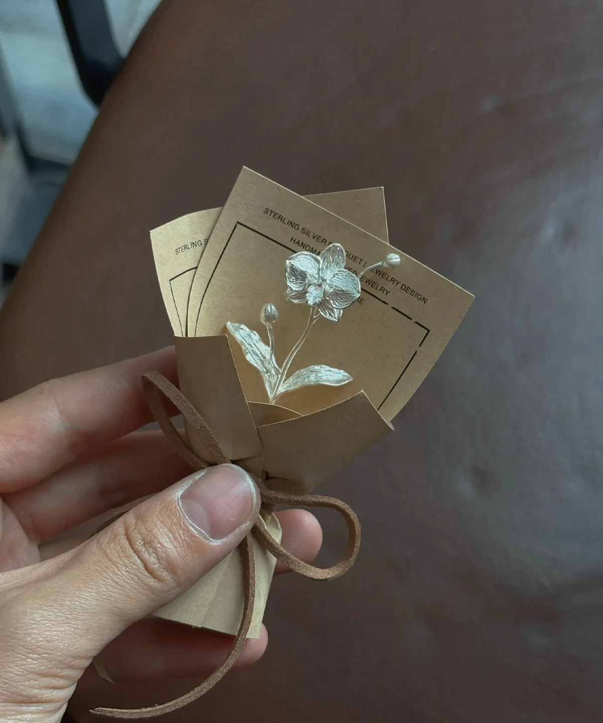Hand holding a 925 sterling silver Phalaenopsis mini bouquet art gift, wrapped in brown paper with suede tie, featuring two delicate handmade silver flower pins on cards, perfect for elegant gifting and floral jewelry lovers.
