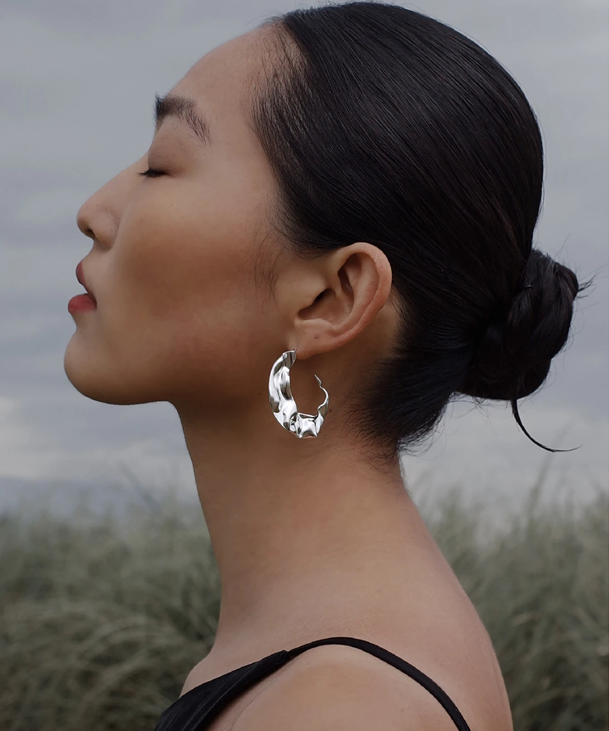 Close-up side profile of a woman wearing Lazesoul’s Moon Aria 990 pure silver sculptural Moon Earrings, set against a serene natural landscape with tall grass and cloudy sky, highlighting the handmade artistry and elegant lunar-inspired design.