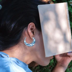 A serene outdoor scene featuring a woman in a light blue shirt lying on grass, reading a book, wearing Lazesoul’s Moon Aria 990 pure silver sculptural Moon Earrings with a textured, luminous design, handmade artisanal jewelry inspired by the moon.