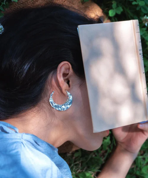 A serene outdoor scene featuring a woman in a light blue shirt lying on grass, reading a book, wearing Lazesoul’s Moon Aria 990 pure silver sculptural Moon Earrings with a textured, luminous design, handmade artisanal jewelry inspired by the moon.