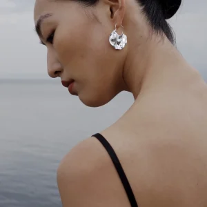 Side profile of a woman by calm water wearing Lazesoul’s Moon Aria – 990 Pure Silver Sculptural Moon Earrings; handmade art jewelry with an abstract lunar design, blending modern elegance with nature‑inspired artistry.
