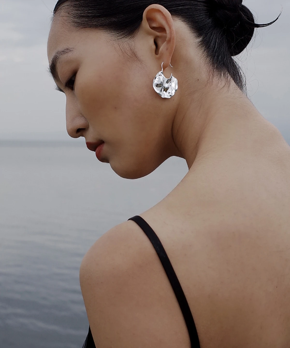 Side profile of a woman by calm water wearing Lazesoul’s Moon Aria – 990 Pure Silver Sculptural Moon Earrings; handmade art jewelry with an abstract lunar design, blending modern elegance with nature‑inspired artistry.
