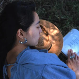 A serene outdoor scene featuring Lazesoul’s Moon Aria-990 Pure Silver Sculptural Moon Earrings, worn by a woman reading on the grass. The handmade art jewelry catches dappled sunlight, highlighting the moon-inspired design’s elegance and craftsmanship.