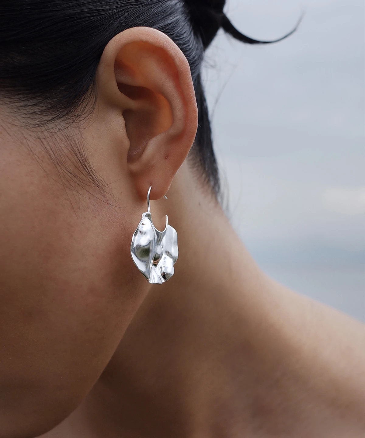 Close-up of Lazesoul’s Moon Aria 990 pure silver sculptural Moon Earrings, handmade with an abstract, organic design. The reflective silver curves catch the light, evoking moonlit elegance against a soft, cloudy sky backdrop.