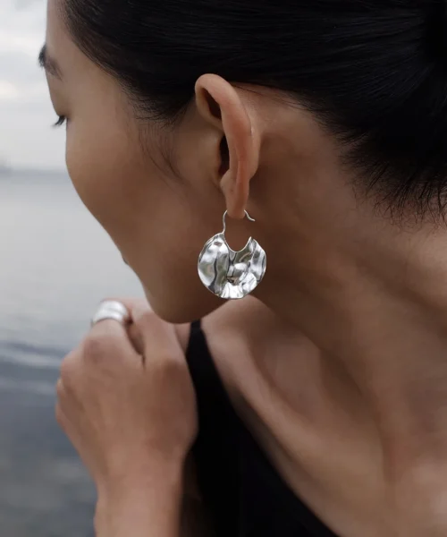 Close-up of Lazesoul Moon Aria 990 pure silver sculptural Moon Earrings, handmade with an organic, crumpled texture. Worn outdoors by the water, the artisanal design reflects moonlit elegance and contemporary art jewelry craftsmanship.