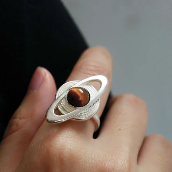 Hand wearing Lazesoul’s handmade Saturn Ring in 925 sterling silver, featuring a sculptural tiger’s eye gemstone as Saturn with orbit-inspired rings; artisanal celestial jewelry with modern elegance.