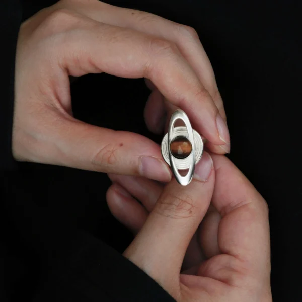 Close-up of hands holding Lazesoul’s handmade Saturn Ring in 925 sterling silver, featuring a polished tiger’s eye gemstone as Saturn with sculptural orbit bands; celestial-inspired artisan jewelry blending cosmic symbolism with timeless elegance.