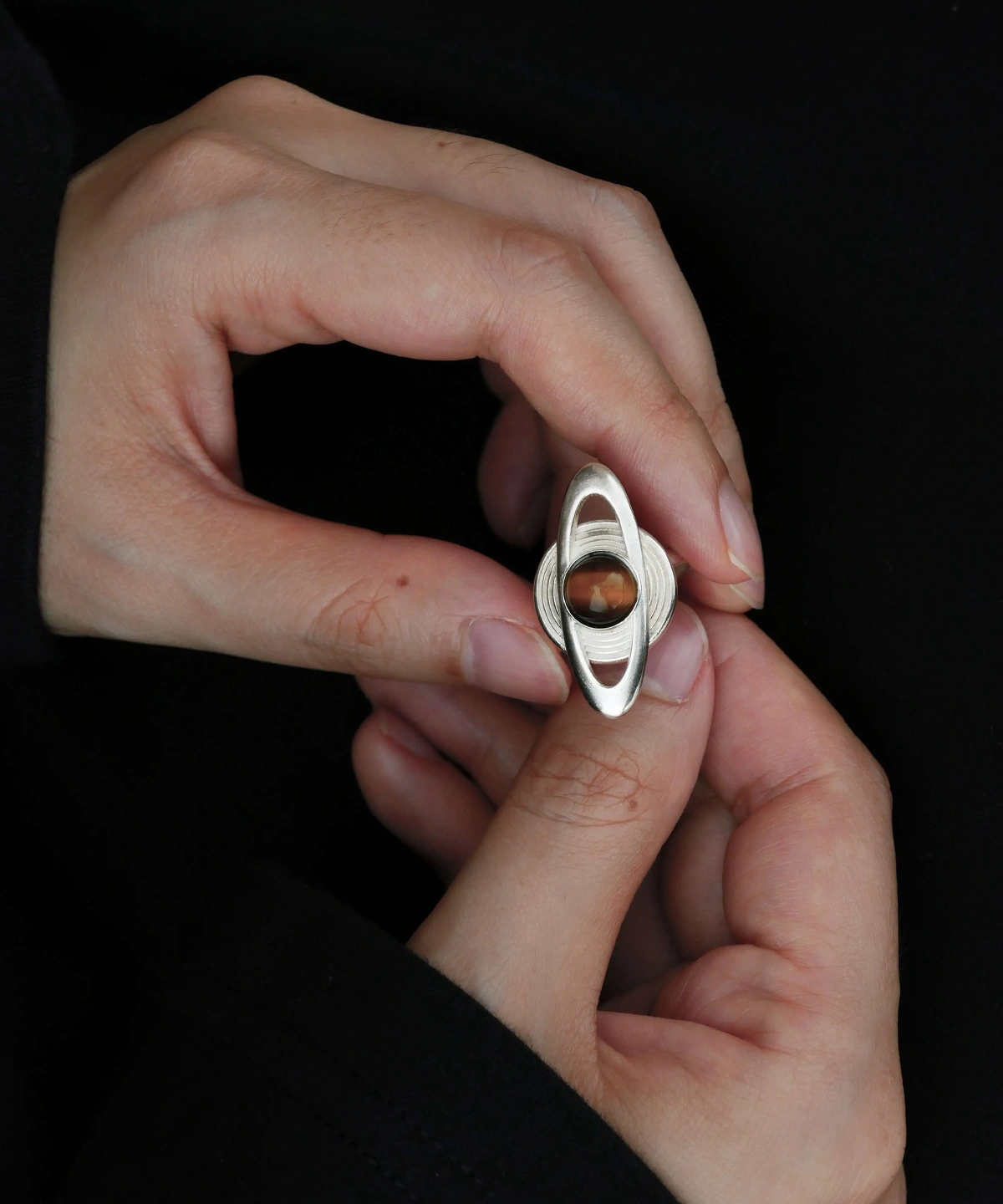 Close-up of hands holding Lazesoul’s handmade Saturn Ring in 925 sterling silver, featuring a polished tiger’s eye gemstone as Saturn with sculptural orbit bands; celestial-inspired artisan jewelry blending cosmic symbolism with timeless elegance.