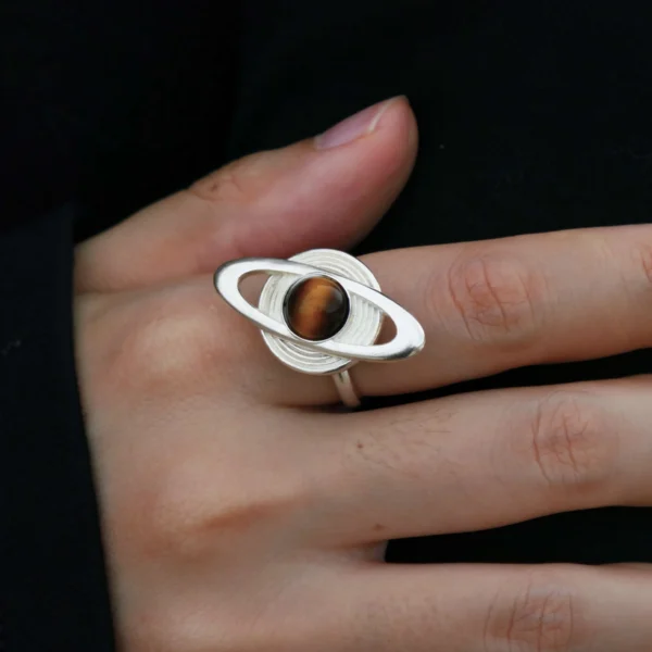 Close-up of hands holding Lazesoul’s handmade Saturn Ring in 925 sterling silver, featuring a polished tiger’s eye gemstone as Saturn with orbit-inspired bands; celestial artisan jewelry blending cosmic symbolism with timeless elegance.