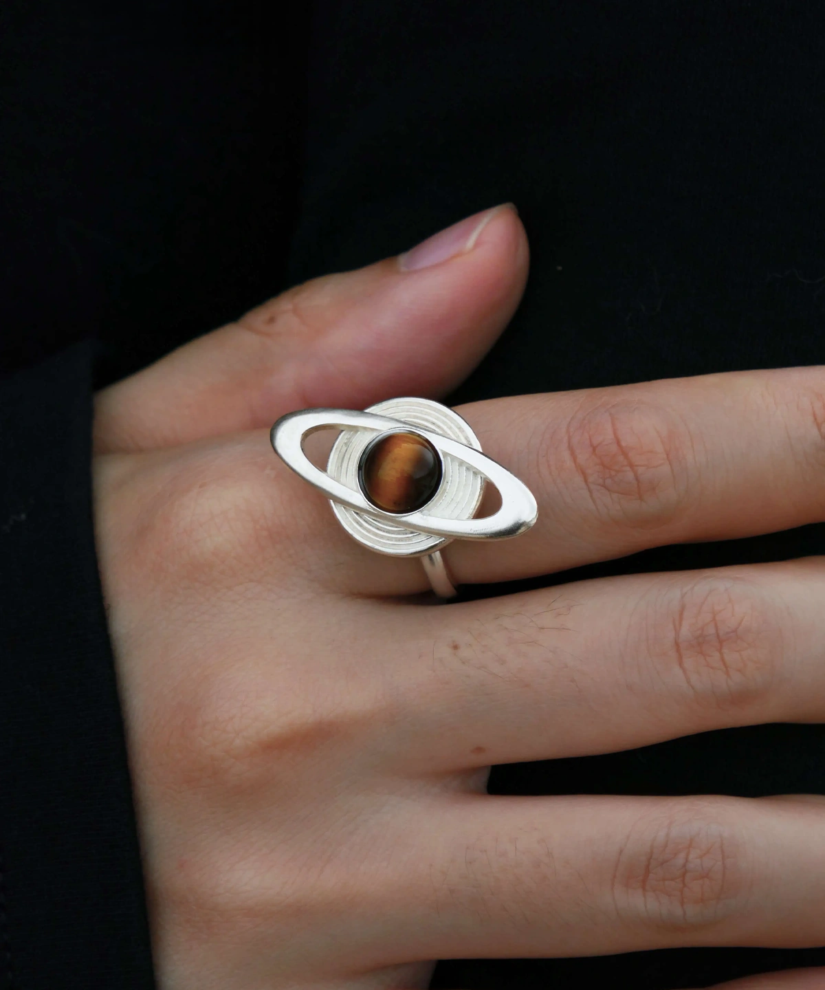 Close-up of hands holding Lazesoul’s handmade Saturn Ring in 925 sterling silver, featuring a polished tiger’s eye gemstone as Saturn with orbit-inspired bands; celestial artisan jewelry blending cosmic symbolism with timeless elegance.