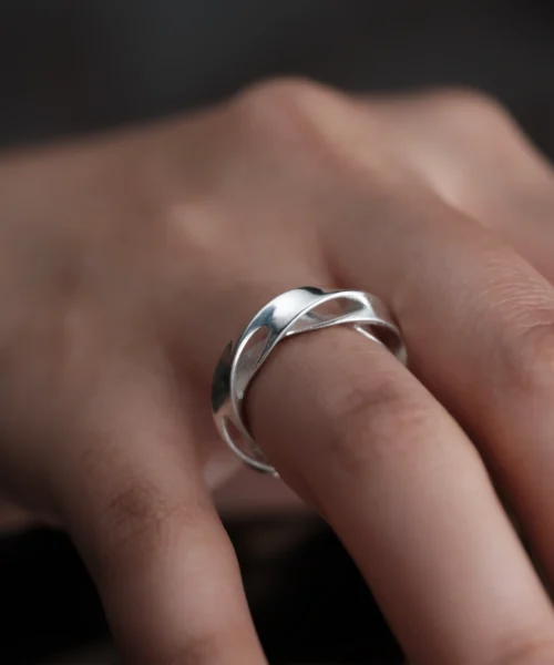 Close-up of a hand wearing Lazesoul Handmade Art Jewelry S925 Sterling Silver Hollow Mobius Strip Ring on the middle finger, featuring two interwoven polished silver bands forming a continuous loop, highlighted against a dark blurred background to emphasize craftsmanship.