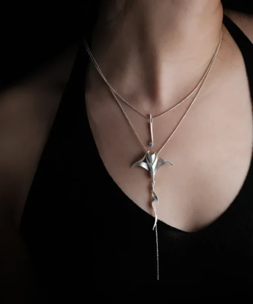 Close-up of a model wearing Lazesoul Handmade Nature Jewelry S925 Sterling Silver Stacked Manta Ray Necklace, featuring a polished manta ray pendant with flowing tail layered alongside a silver bar pendant with blue gemstone, highlighted against a black sleeveless top.