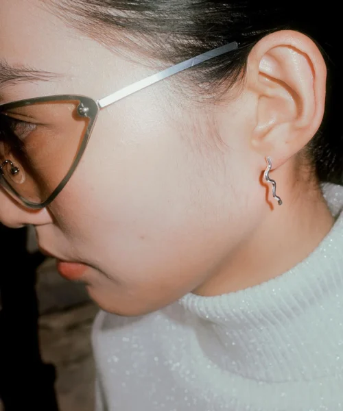 Side profile of a model wearing Lazesoul Handmade Art Jewelry Songs on the Road-Original Design 990 Sterling Silver Fun Earrings | Flute – Nocturne of Warmth, featuring a delicate wavy silver design paired with bold eyeglasses and a white textured turtleneck.