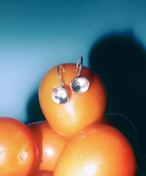 Close-up of Lazesoul Handmade Art Jewelry Songs on the Road-Original Design 990 Sterling Silver Fun Earrings | Trumpet – Overture of Hope, shown as circular silver earrings placed on orange tomatoes arranged like a playful face, highlighted against a blue gradient background.