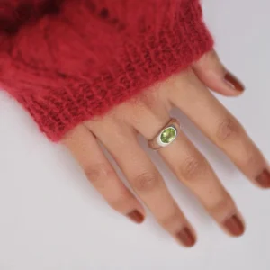 Close-up of a hand wearing a Silver Ring with an oval natural peridot gemstone, styled with brown nail polish and a red knit sweater; elegant August Birthstone Ring showcasing vibrant green hues and handcrafted Natural Peridot Ring artistry.
