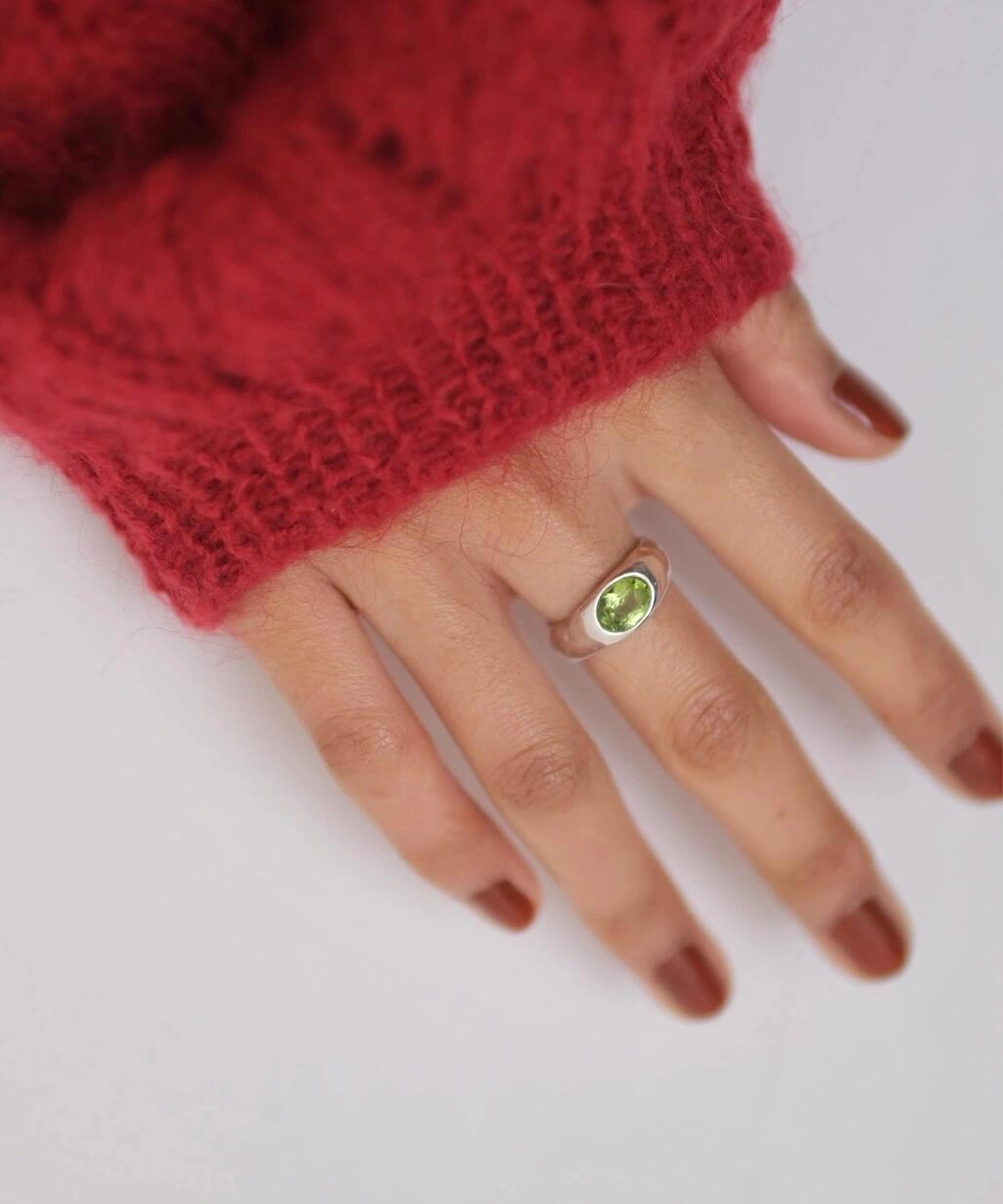 Close-up of a hand wearing a Silver Ring with an oval natural peridot gemstone, styled with brown nail polish and a red knit sweater; elegant August Birthstone Ring showcasing vibrant green hues and handcrafted Natural Peridot Ring artistry.