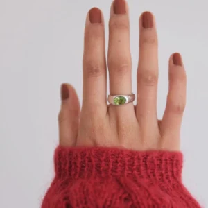 Close-up of a hand wearing a Silver Ring with an oval natural peridot gemstone, styled with brown nail polish and a red knit sweater; elegant August Birthstone Ring showcasing vibrant green hues and handcrafted sterling silver artistry.