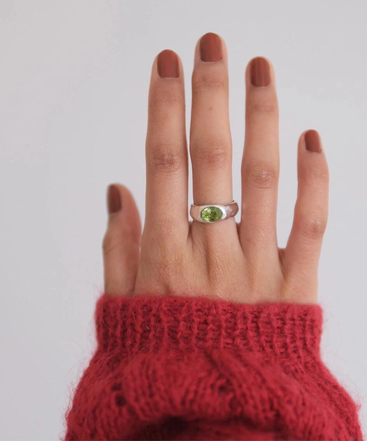 Close-up of a hand wearing a Silver Ring with an oval natural peridot gemstone, styled with brown nail polish and a red knit sweater; elegant August Birthstone Ring showcasing vibrant green hues and handcrafted sterling silver artistry.
