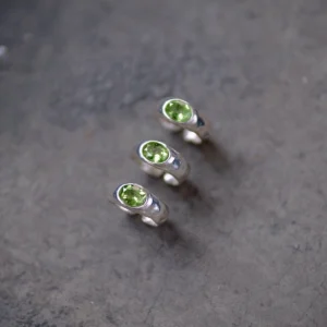 Three handcrafted Silver Rings with round natural peridot gemstones arranged diagonally on a textured gray surface; elegant August Birthstone Ring designs showcasing vibrant green stones and minimalist sterling silver craftsmanship.