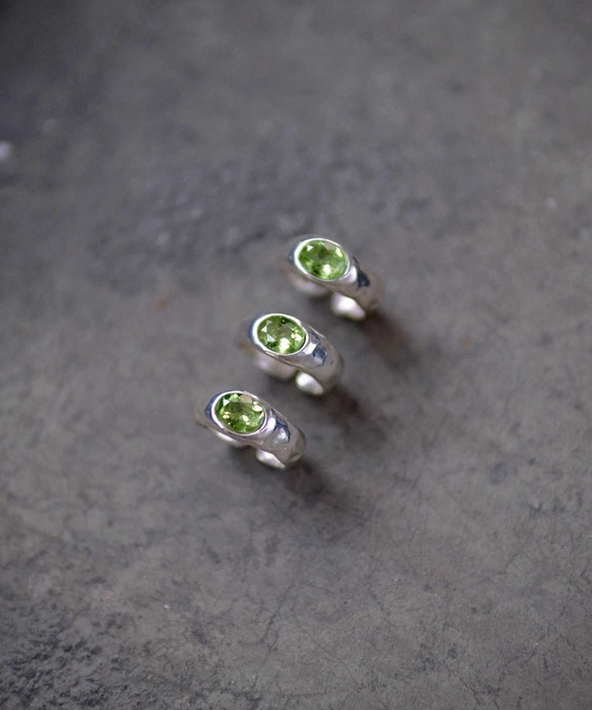 Three handcrafted Silver Rings with round natural peridot gemstones arranged diagonally on a textured gray surface; elegant August Birthstone Ring designs showcasing vibrant green stones and minimalist sterling silver craftsmanship.