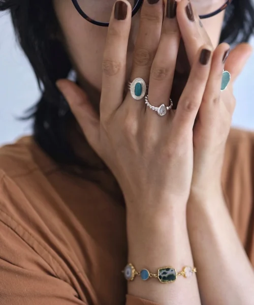 Hands with dark brown nails wearing multiple rings, including a December Birthstone Ring with natural turquoise, styled with a colorful gemstone bracelet and earthy tones.