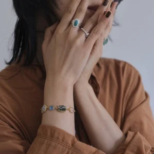 December Birthstone Ring-Natural Turquoise Ring in Sterling Silver 07 Hands with dark nails wearing sterling silver rings, including a December Birthstone Ring with natural turquoise, styled with a gold bracelet featuring multicolored stones and a brown shirt.