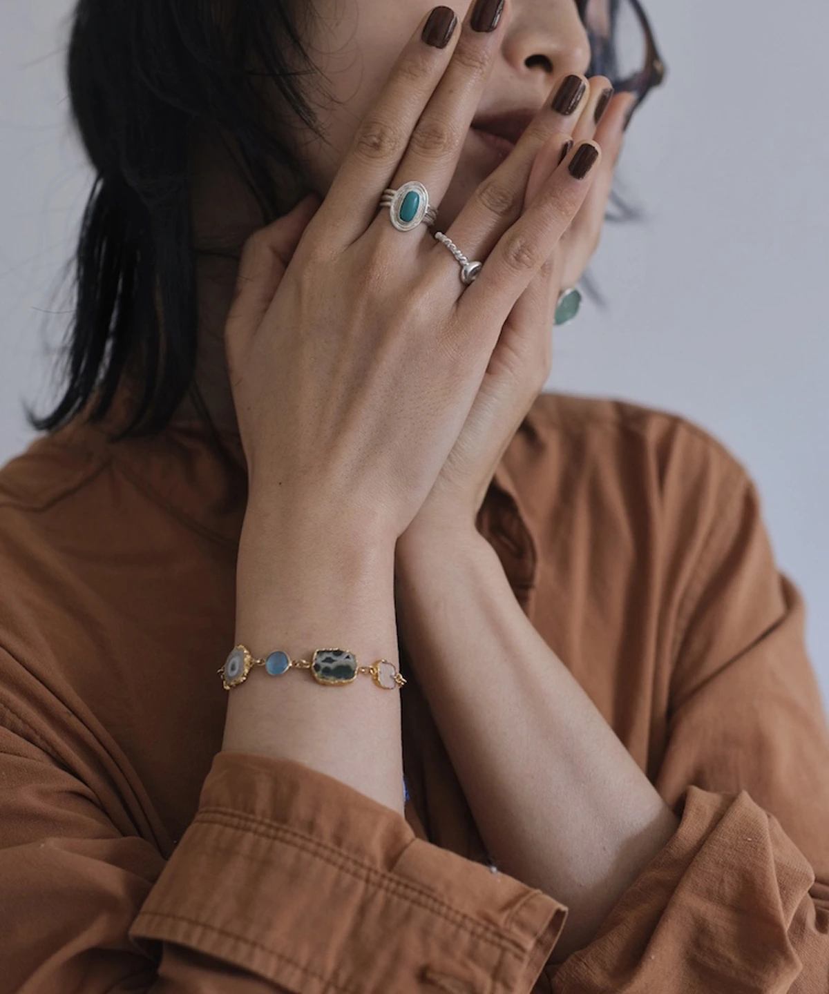 December Birthstone Ring-Natural Turquoise Ring in Sterling Silver 07 Hands with dark nails wearing sterling silver rings, including a December Birthstone Ring with natural turquoise, styled with a gold bracelet featuring multicolored stones and a brown shirt.