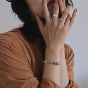 December Birthstone Ring-Natural Turquoise Ring in Sterling Silver 08 Close-up of hands near face wearing sterling silver rings, including a December Birthstone Ring with natural turquoise, styled with a gold bracelet featuring blue and green stones.
