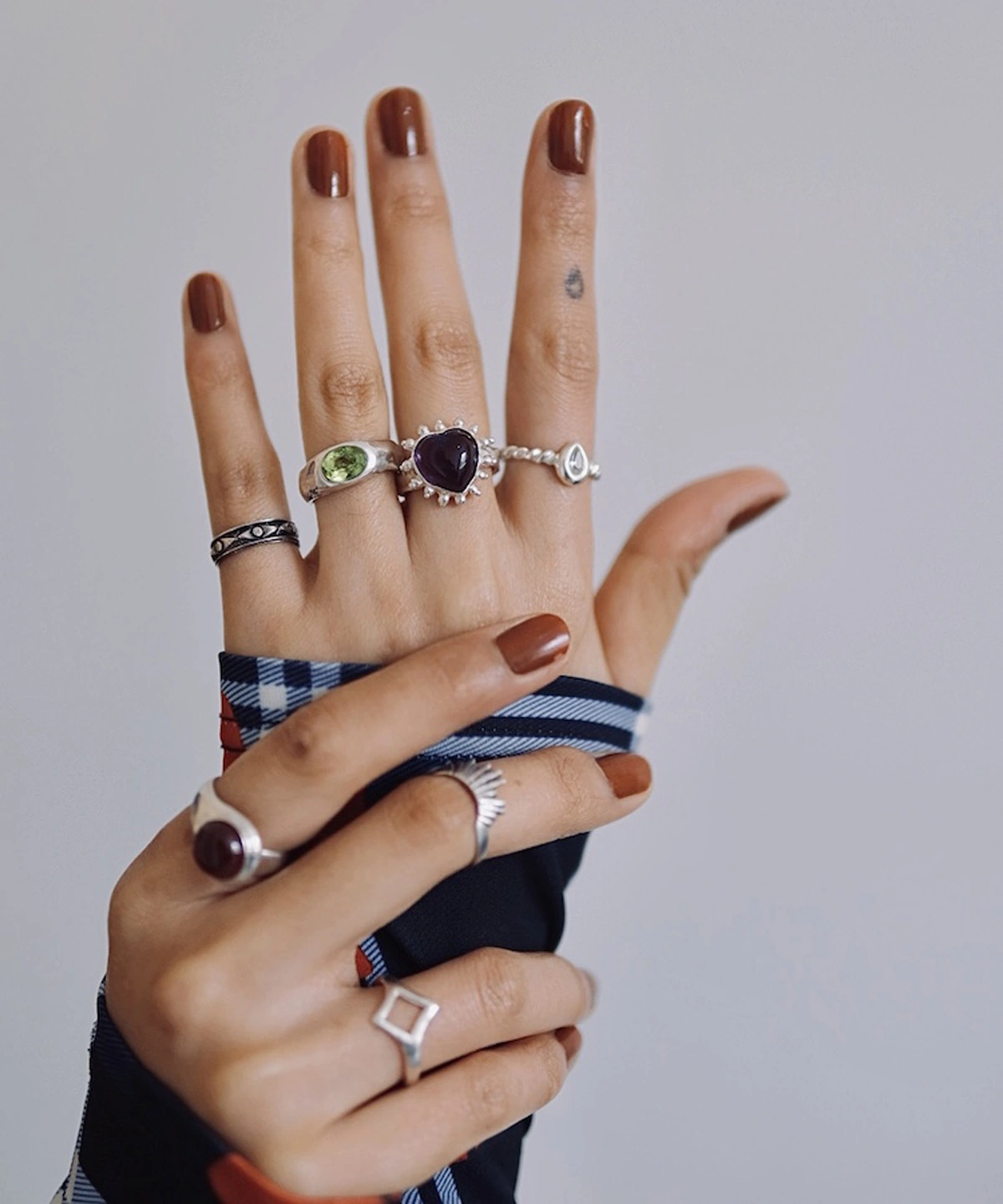 February Birthstone Ring-Natural Amethyst Ring in Sterling Silver 02 Two hands with brown nails wearing multiple silver rings, including a February Birthstone Ring with heart-shaped natural amethyst, styled with teardrop, sunburst, and geometric accents.