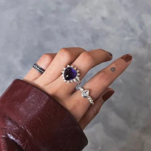 February Birthstone Ring-Natural Amethyst Ring in Sterling Silver 03 Hand with brown nails wearing silver rings, including a February Birthstone Ring with heart-shaped natural amethyst, styled with teardrop and engraved bands against a gray background.