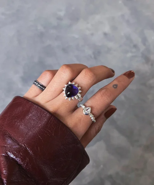 Hand with brown nails wearing silver rings, including a February Birthstone Ring with heart-shaped natural amethyst, styled with teardrop and engraved bands against a gray background.