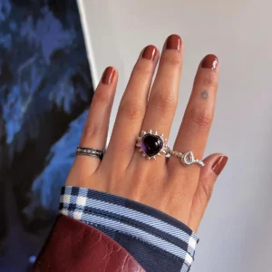 February Birthstone Ring-Natural Amethyst Ring in Sterling Silver 04 Hand with brown nails wearing silver rings, including a February Birthstone Ring with heart-shaped natural amethyst, styled with teardrop and engraved bands against a mixed background.