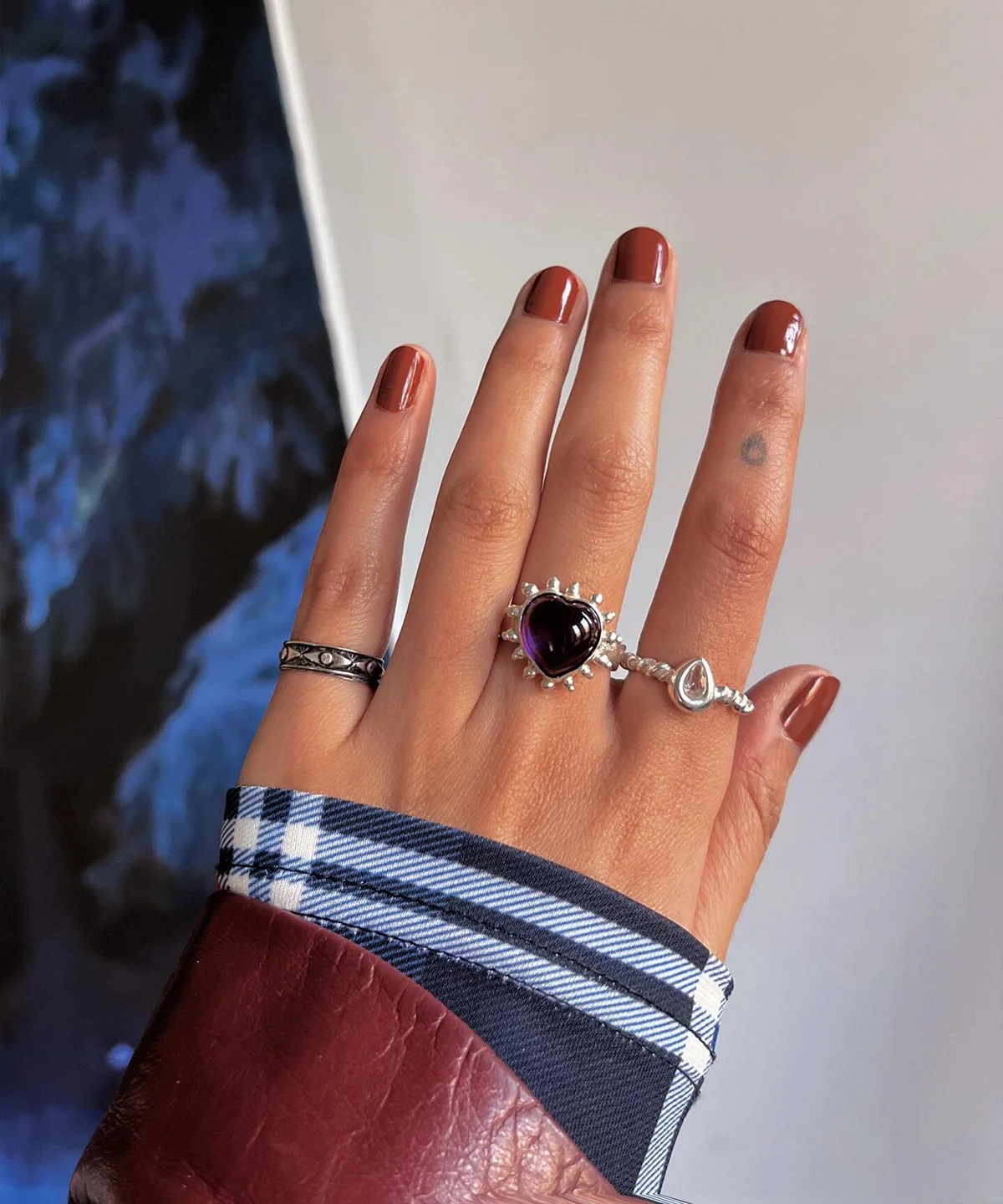 February Birthstone Ring-Natural Amethyst Ring in Sterling Silver 04 Hand with brown nails wearing silver rings, including a February Birthstone Ring with heart-shaped natural amethyst, styled with teardrop and engraved bands against a mixed background.