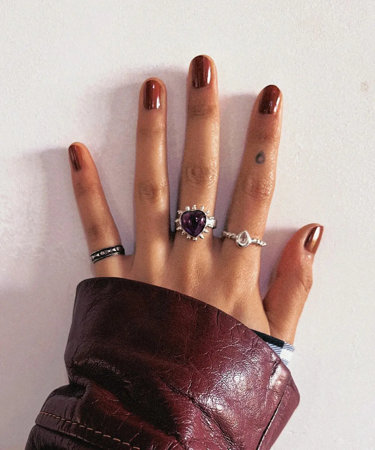 February Birthstone Ring-Natural Amethyst Ring in Sterling Silver 05 Hand with brown nails wearing multiple rings, including a February Birthstone Ring with heart-shaped natural amethyst in sterling silver, styled with teardrop and engraved bands.