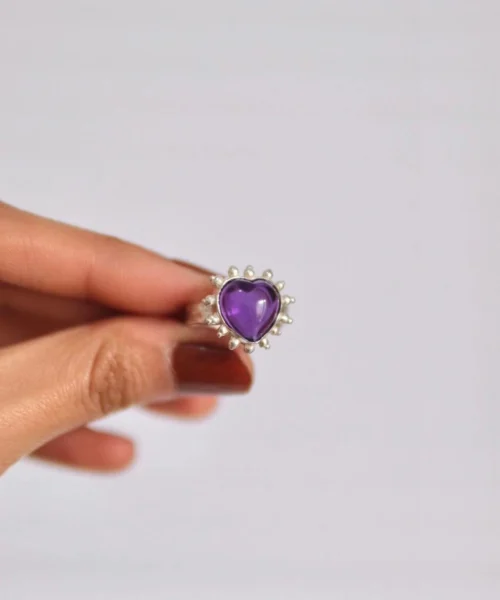 Hand holding a silver ring with a heart-shaped natural amethyst gemstone, surrounded by beaded detailing, showcasing the February Birthstone Ring in sterling silver.