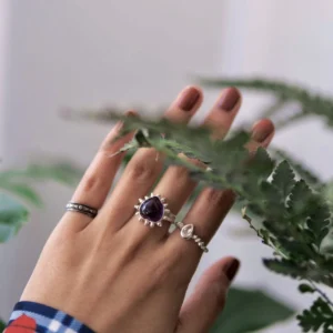 February Birthstone Ring-Natural Amethyst Ring in Sterling Silver 07 Hand with muted brown nails wearing three silver rings, including a February Birthstone Ring with natural amethyst, partially obscured by fern leaves and styled with a plaid shirt.