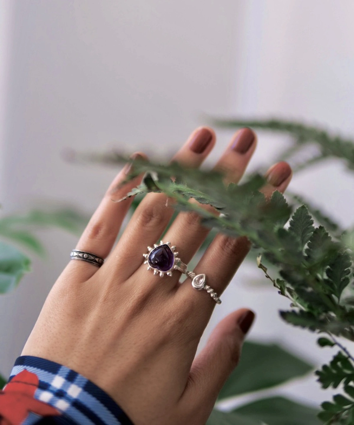 February Birthstone Ring-Natural Amethyst Ring in Sterling Silver 07 Hand with muted brown nails wearing three silver rings, including a February Birthstone Ring with natural amethyst, partially obscured by fern leaves and styled with a plaid shirt.