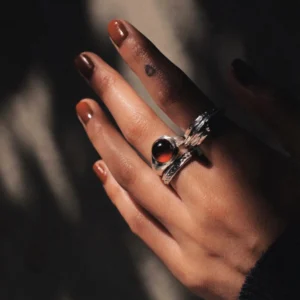 Hand with brown nails wearing sterling silver rings, including a January Birthstone Ring with natural garnet, styled with leaf-inspired silver band and circular tattoo.