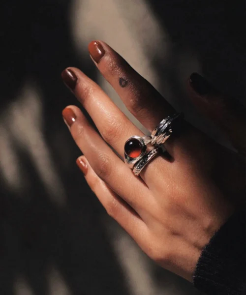 Hand with brown nails wearing sterling silver rings, including a January Birthstone Ring with natural garnet, styled with leaf-inspired silver band and circular tattoo.